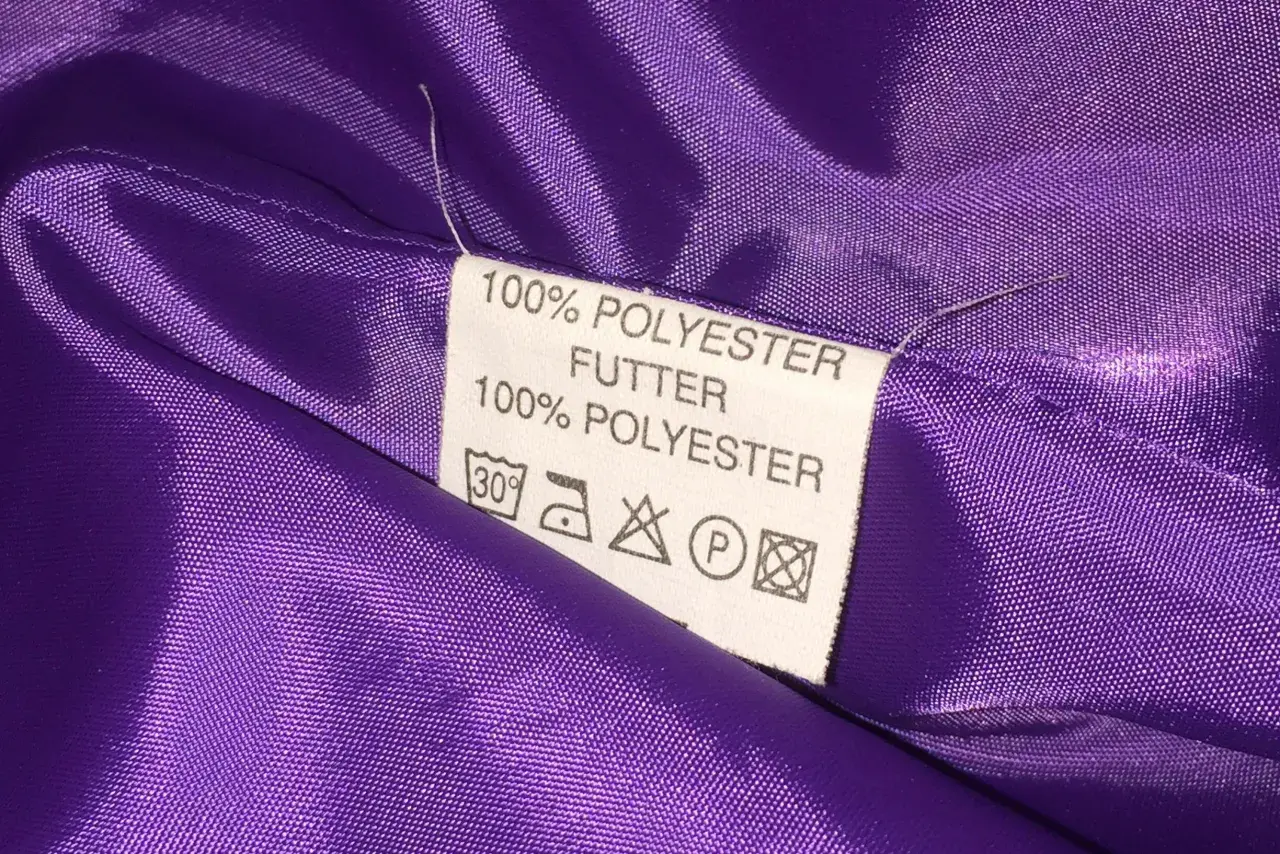 Metka z informacją "100% POLIESTER FUTTER" na fioletowej, błyszczącej tkaninie. Czy poliester jest zdrowy? Ta metka nie odpowie na to pytanie.